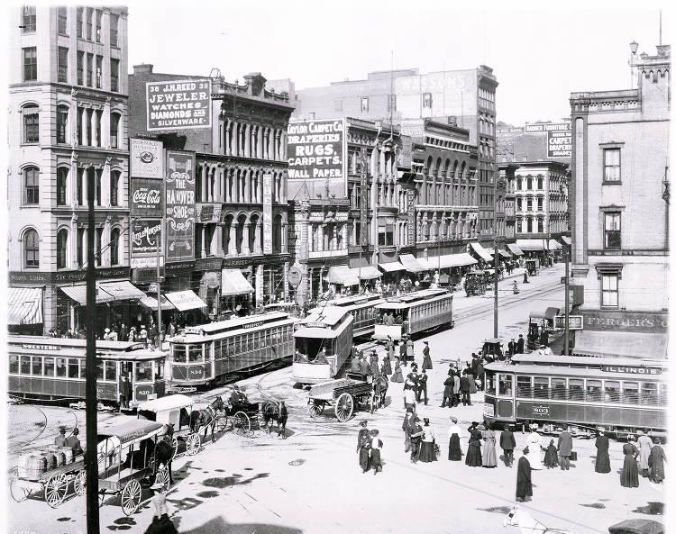 A busy intersection in the middle of a city. The intersection is filled with several streetcars, people, and horse-drawn wagons.