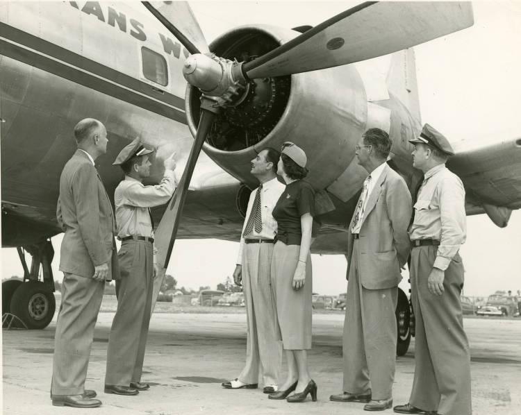 Two pilots, a flight attendant and three men stand looking at an airplane's propeller.