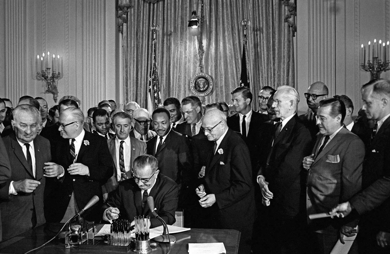 President Johnson sits at a desk and signs a document. A large group of people stand behind him watching.