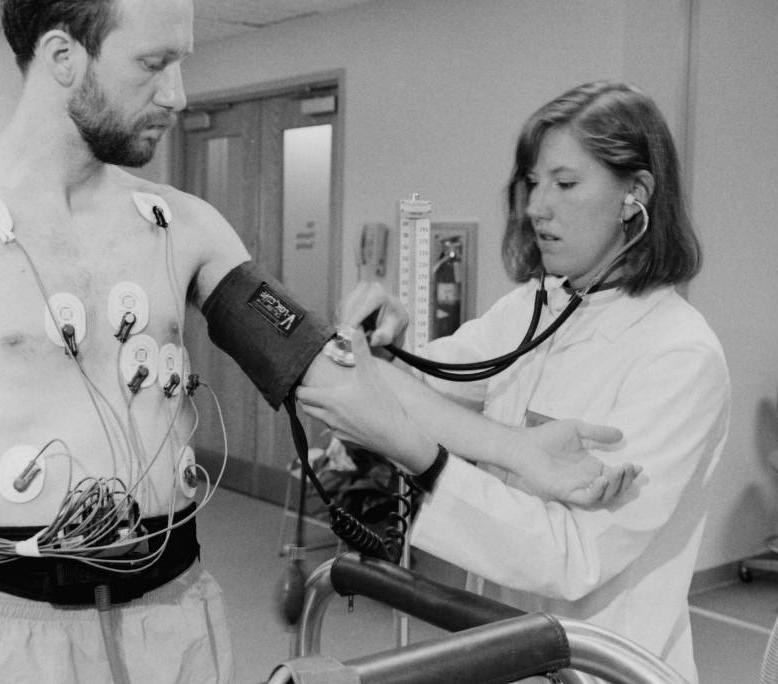 A doctor takes a man's blood pressure using a cuff. The man has several wires attached to his body to monitor vitals. 