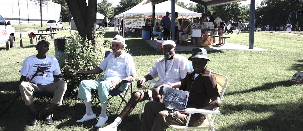 A group of four men sit in lawn chairs under a tree.