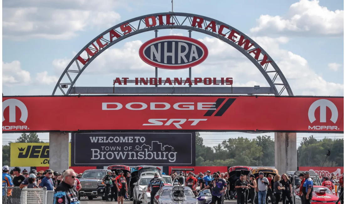 A crowd of people and cars are in front of an arched sign. In the top of the arch is written "Lucas Oil Raceway". Under that is "NHRA At Indianapolis". A long, red, rectangular sign stretches across the middle of the archway and reads "Dodge SRT". Below that is a sign reading "Welcome to Town of Brownsburg"