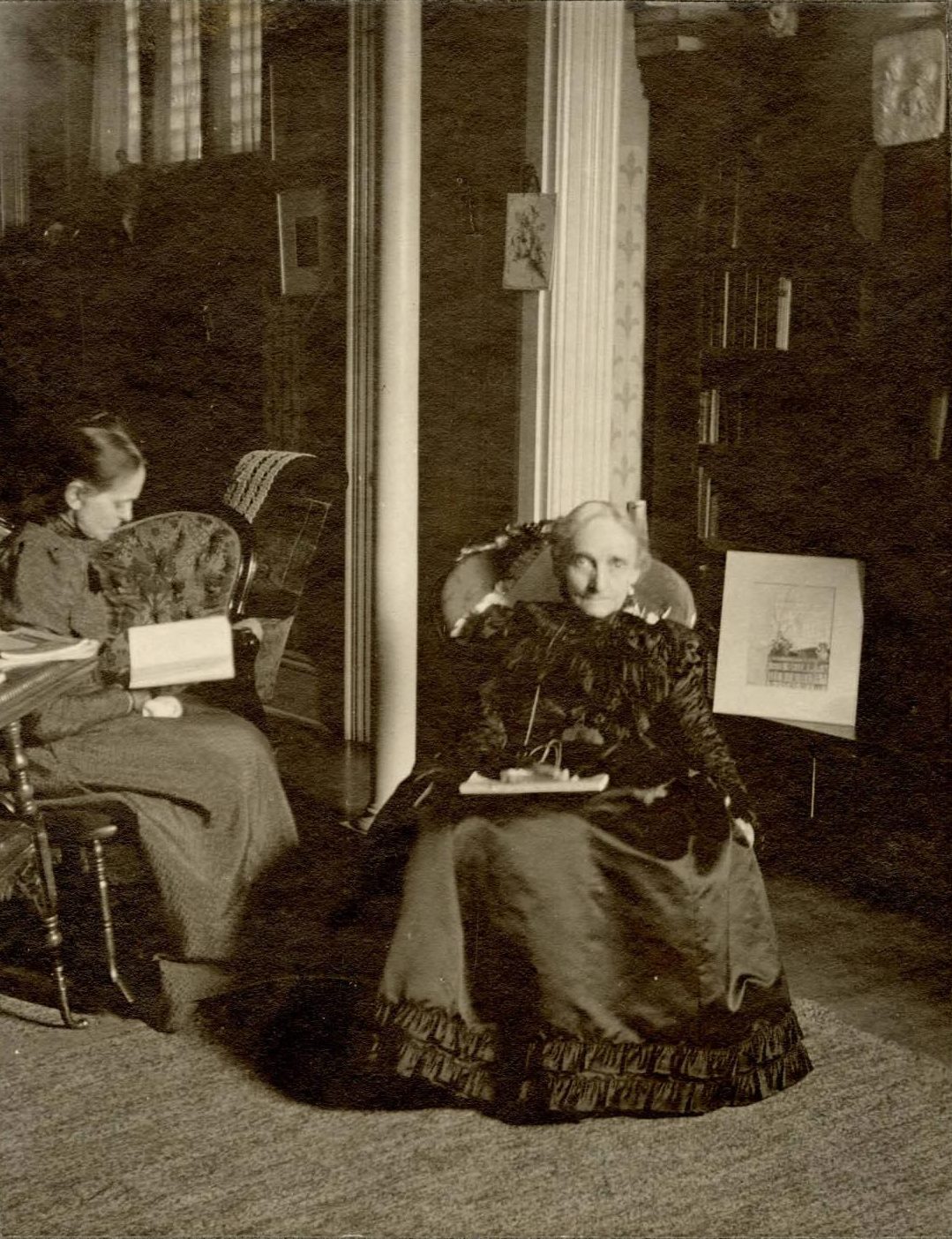 Catherine Merrill is seated next to her sister wearing a dark dress with a full skirt.