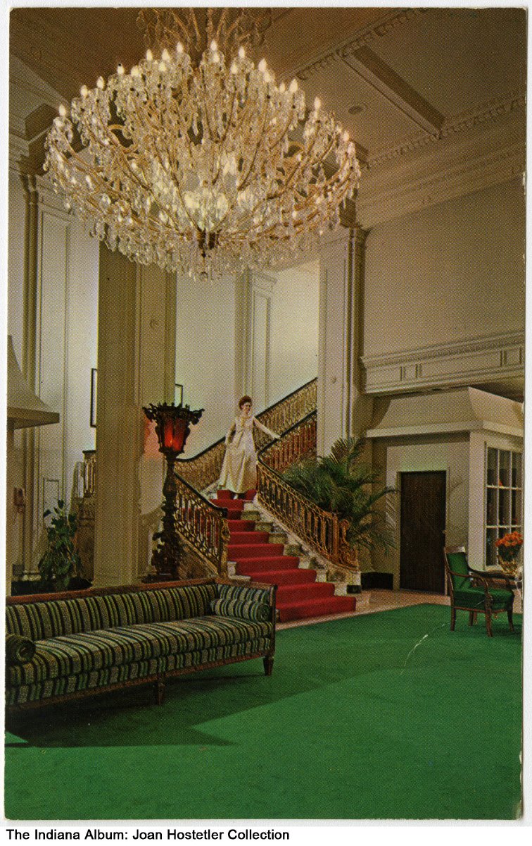 A woman in a ball gown descends from a carpeted staircase into a large room with a giant chandelier. 