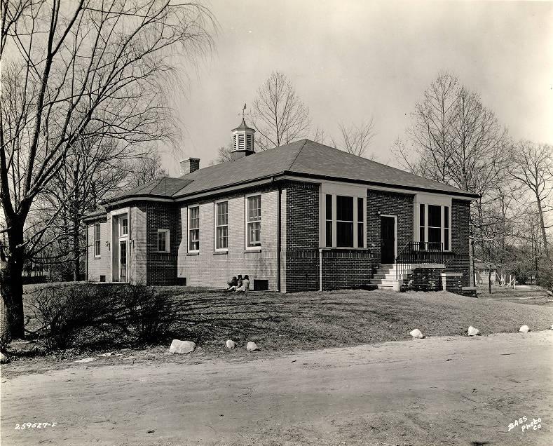 Three children sit in the grass outside of a small brick building.