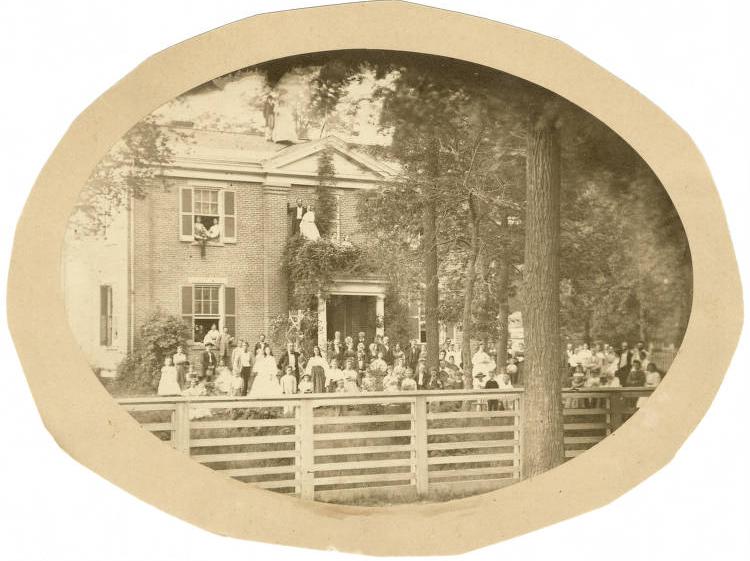 An old sepia photograph shows a large gathering of people in front of a large, brick home.