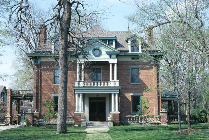 The square, classical brick home has a two-tiered portico entrance supported on each level by white columns. There is covered, brick porch on the right of the house and a porte cochere on the left. The roof is set with dormer windows.