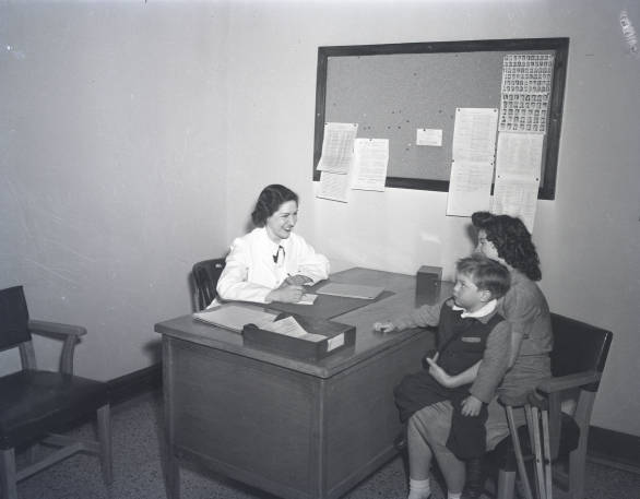 A woman sits behind a desk. She is talking with another woman who is holding a small child. 