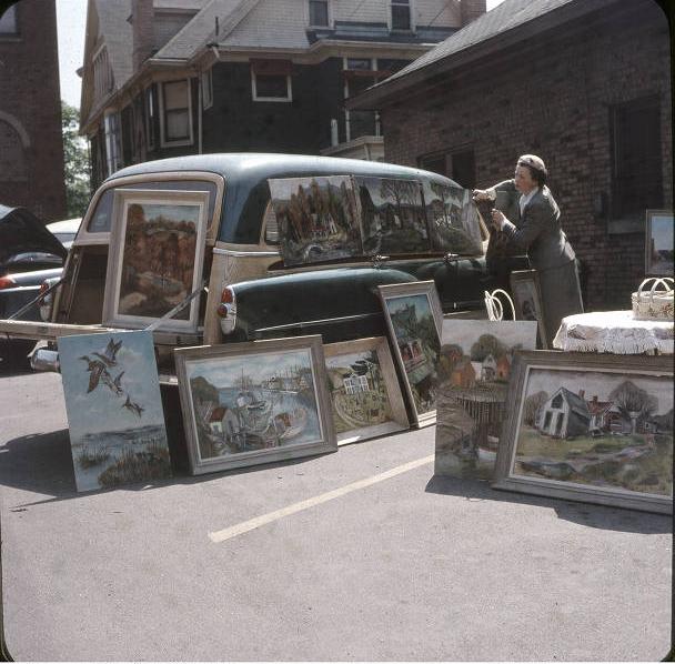 A woman is placing paintings on and around a car in a parking lot.