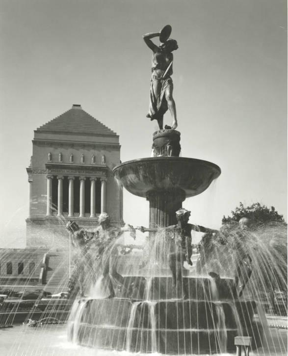 A fountain made up of bronze figures that create five tiers around a granite base. In the background is the Indiana World War Memorial.