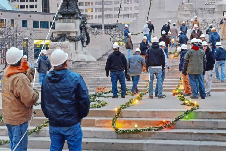 A group of people stand on the steps of the Soldiers and Sailors Monument. Garland with lights lay on the ground. 