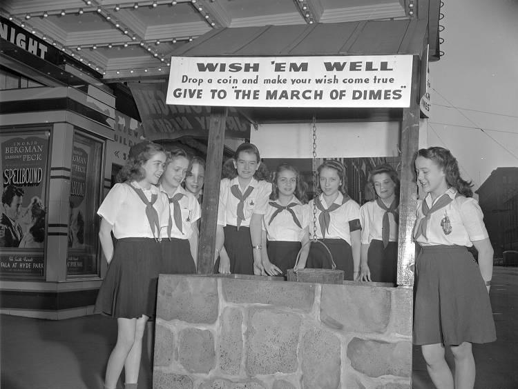 A group of young women stand around a well with a sign inviting people to donate change. 