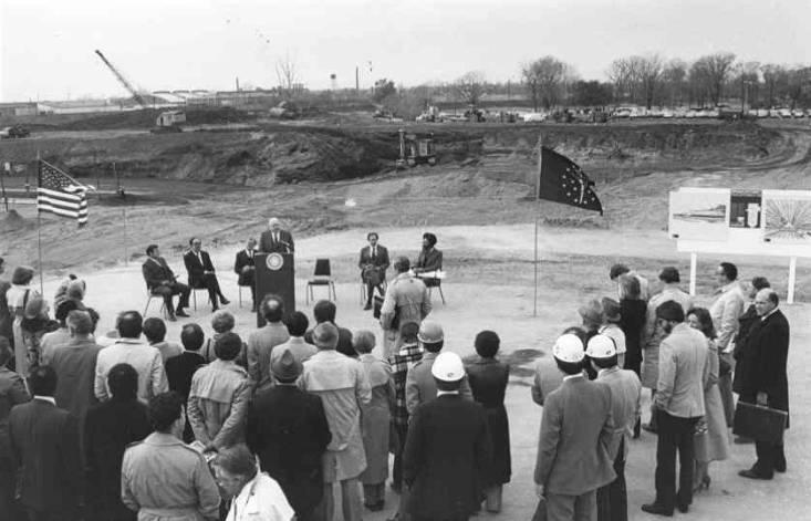 A crowd gathers to hear a presentation of a man standing at a podium in the middle of cleared land. 