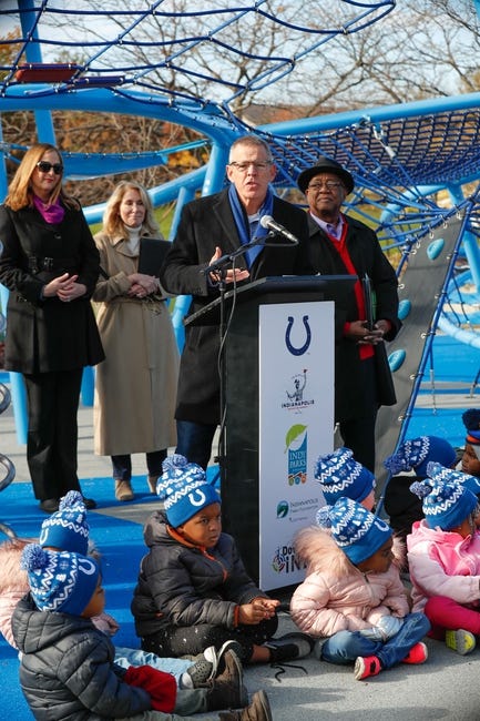 A man stands behind a podium. Several people stand behind him and children sit at the base of the podium. Playground equipment is in the background. 
