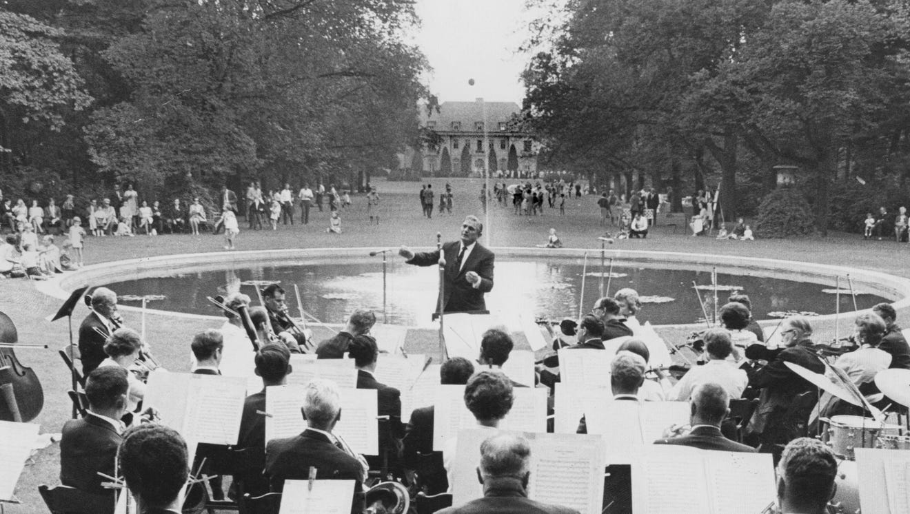 A view looking out from an orchestra towards the conductor. Behind the conductor is a pool of water, a large lawn, and a house. 