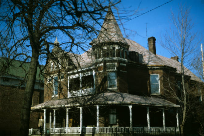 This three-story brick Victorian gothic house has a wrap-around porch and a turret with a conical roof at one corner of the roof and a gable at the other corner. the front door.