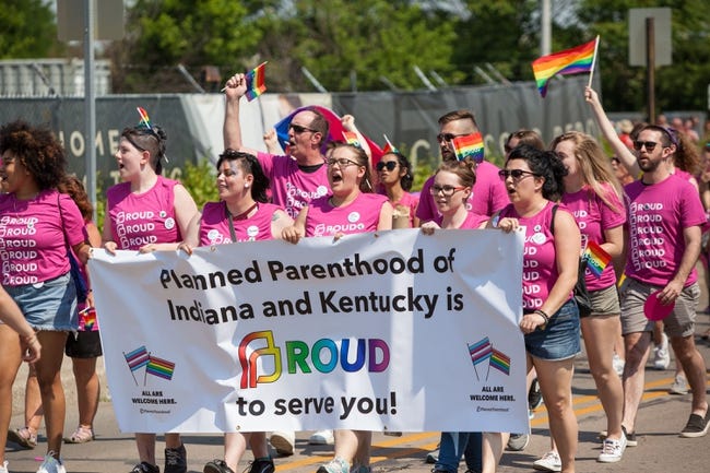 A group of women and men march together. All people are wearing pink Planned Parenthood shirts. The people at the front are carrying a Planned Parenthood banner.