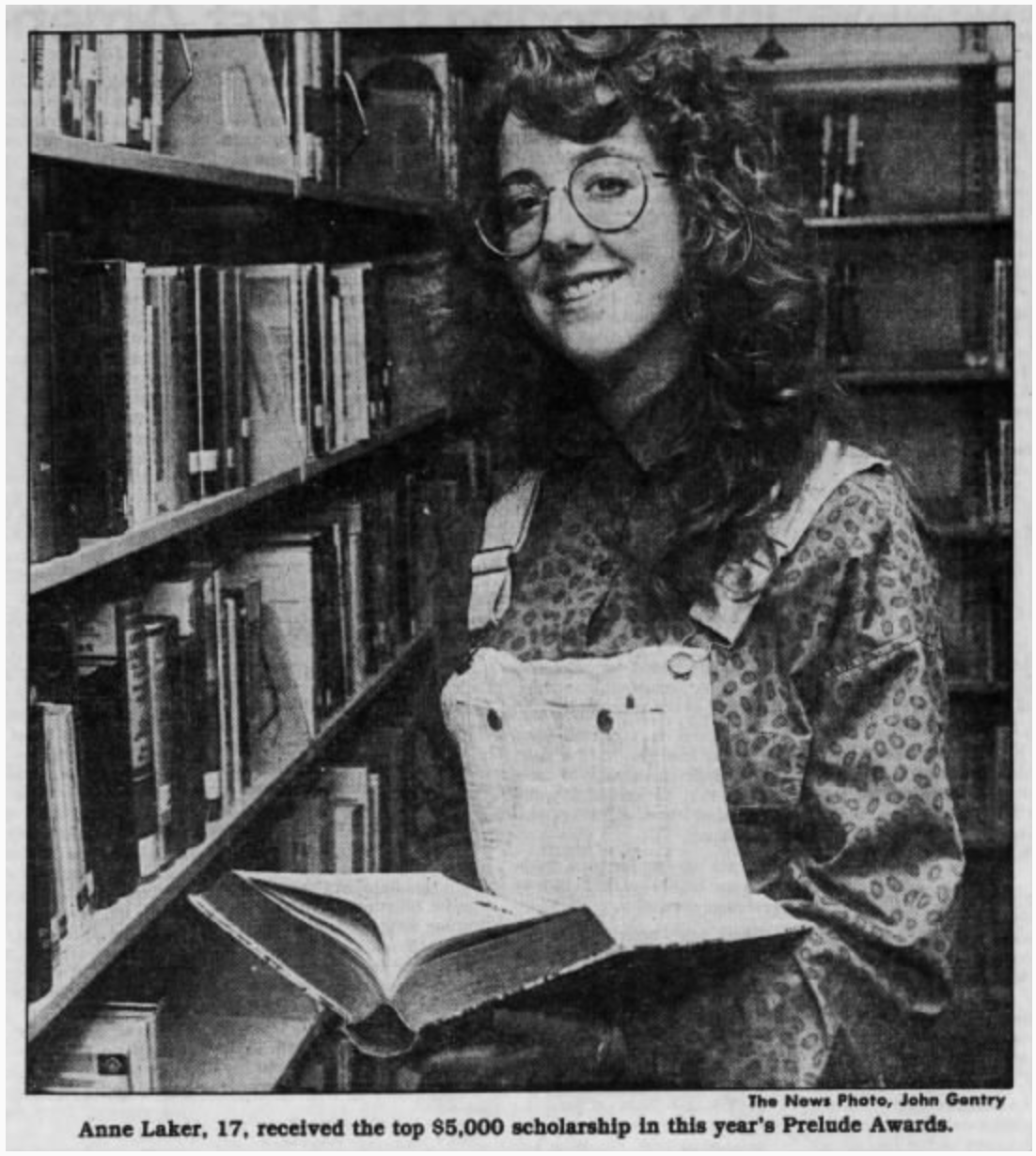 A woman stands in front of a book shelf. She is holding an open book and smiling at the camera.