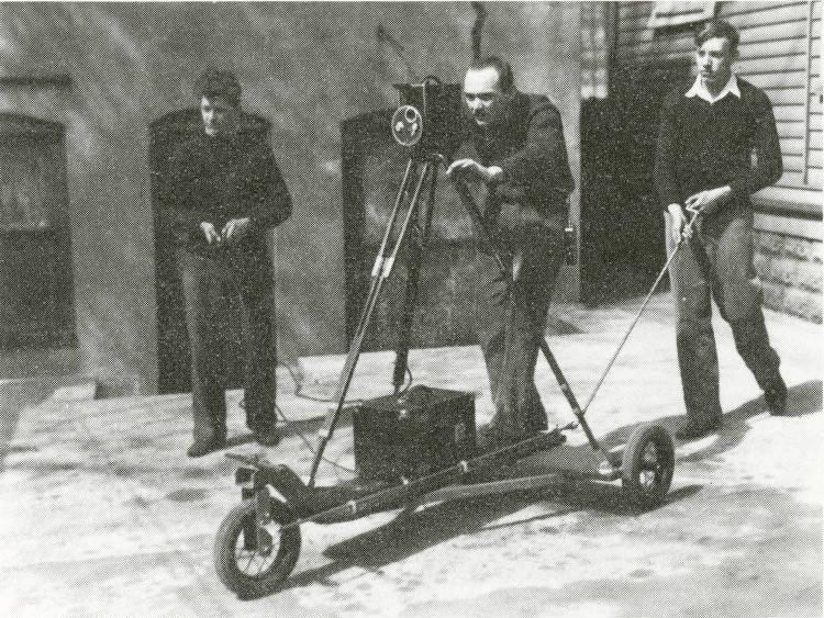 Robert Annis stands behind a camera on a tripod that is attached to a set of wheels. 