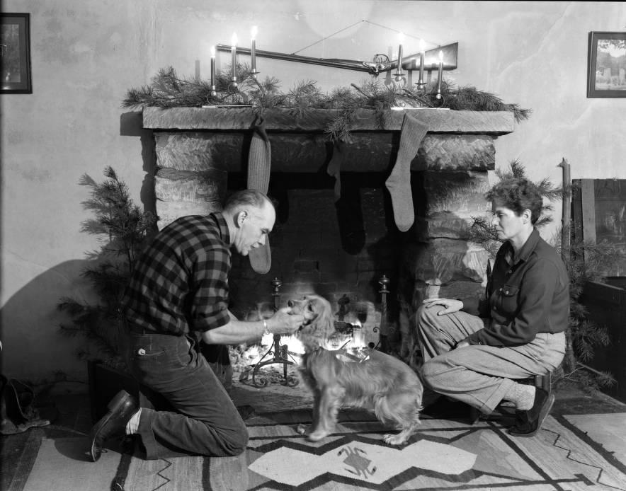 A man and a woman kneel on the floor in front of a fireplace that is decorated for Christmas. The man is petting a dog.  