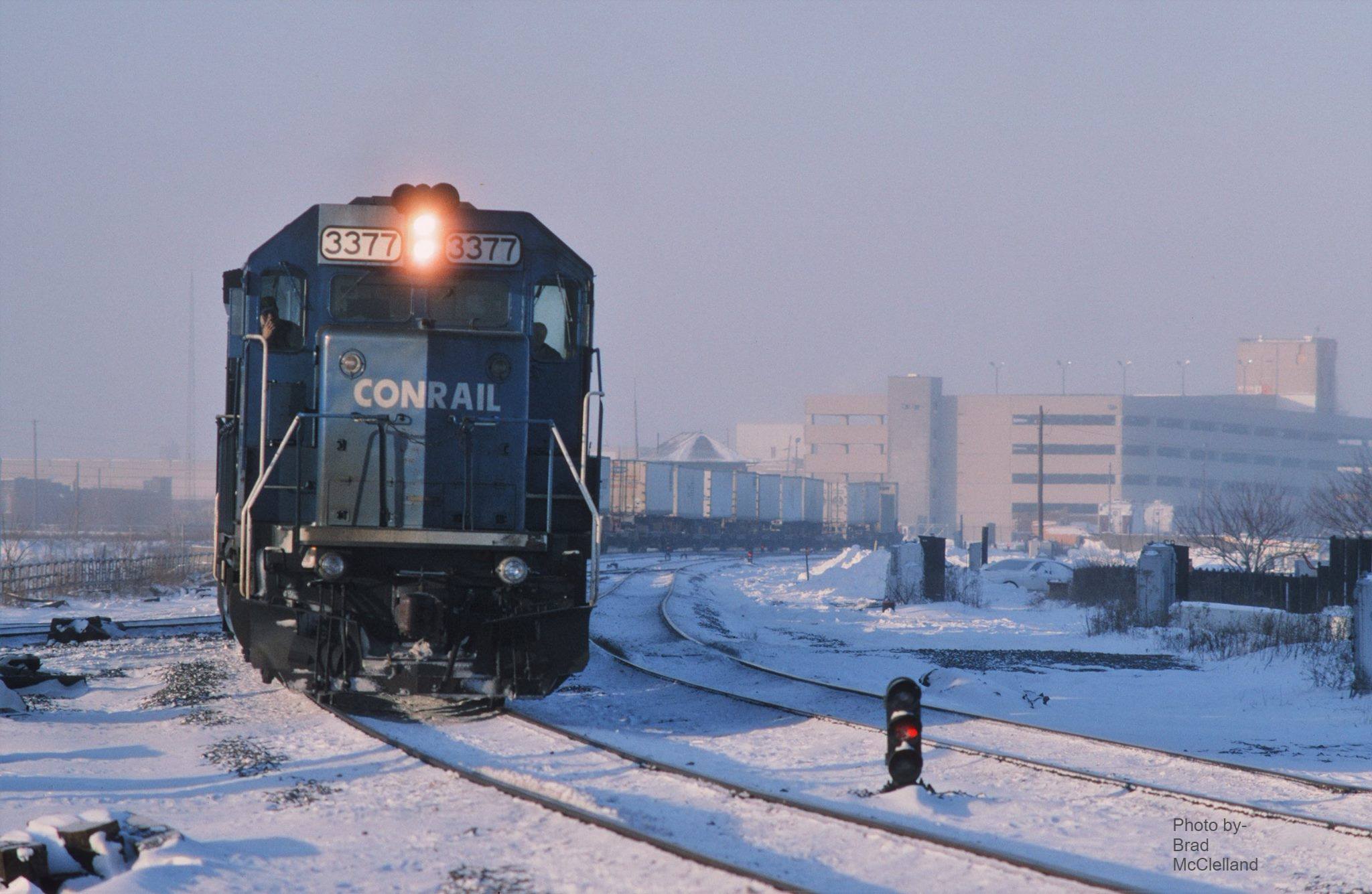 A train runs down snow-covered tracks. The city is in the background.