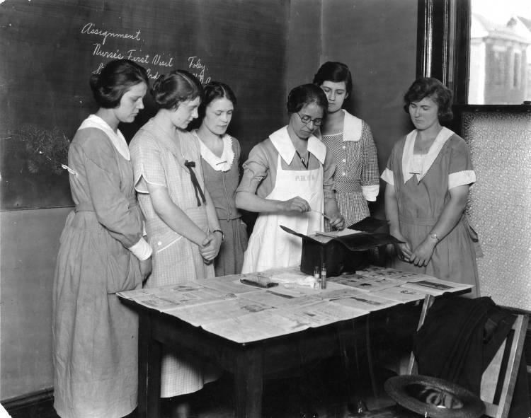 A group of young woman watch as an instructor holds what appears to be a thermometer. 