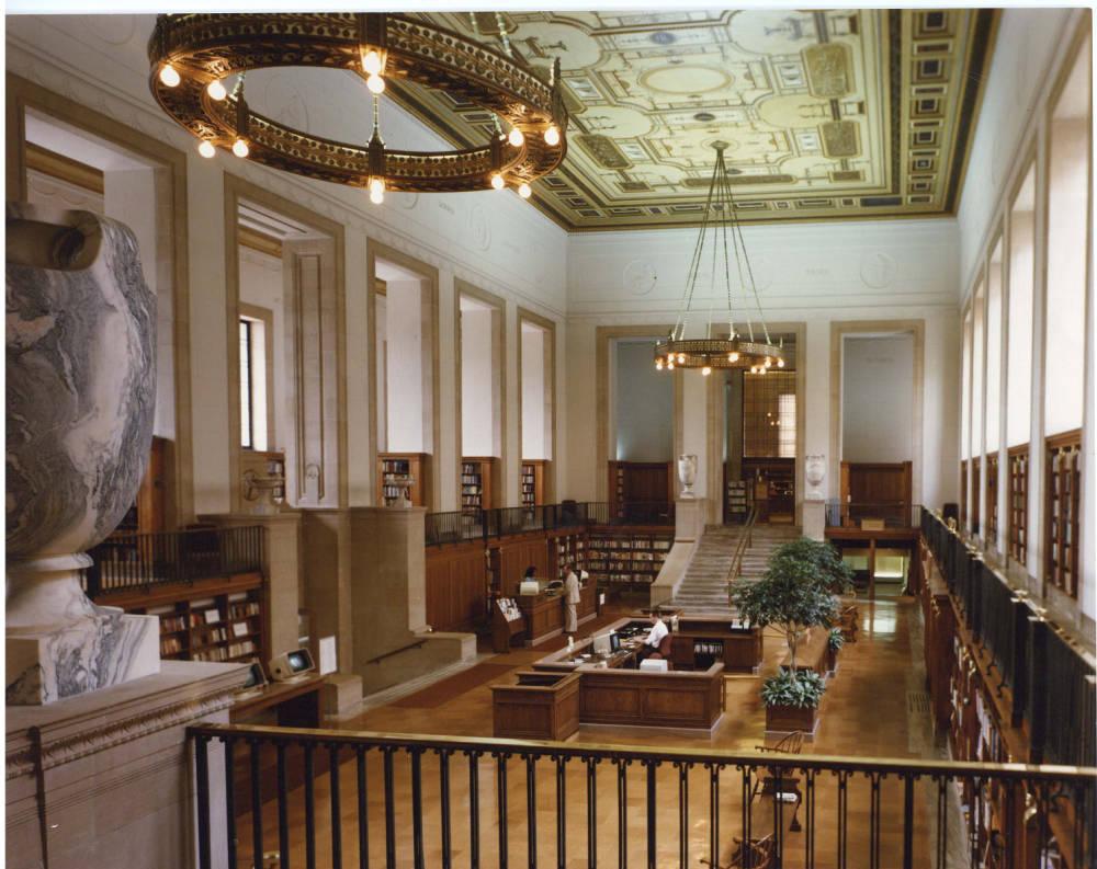 View from an upper floor balcony looking down into a large room with book-lined walls and a central desk.