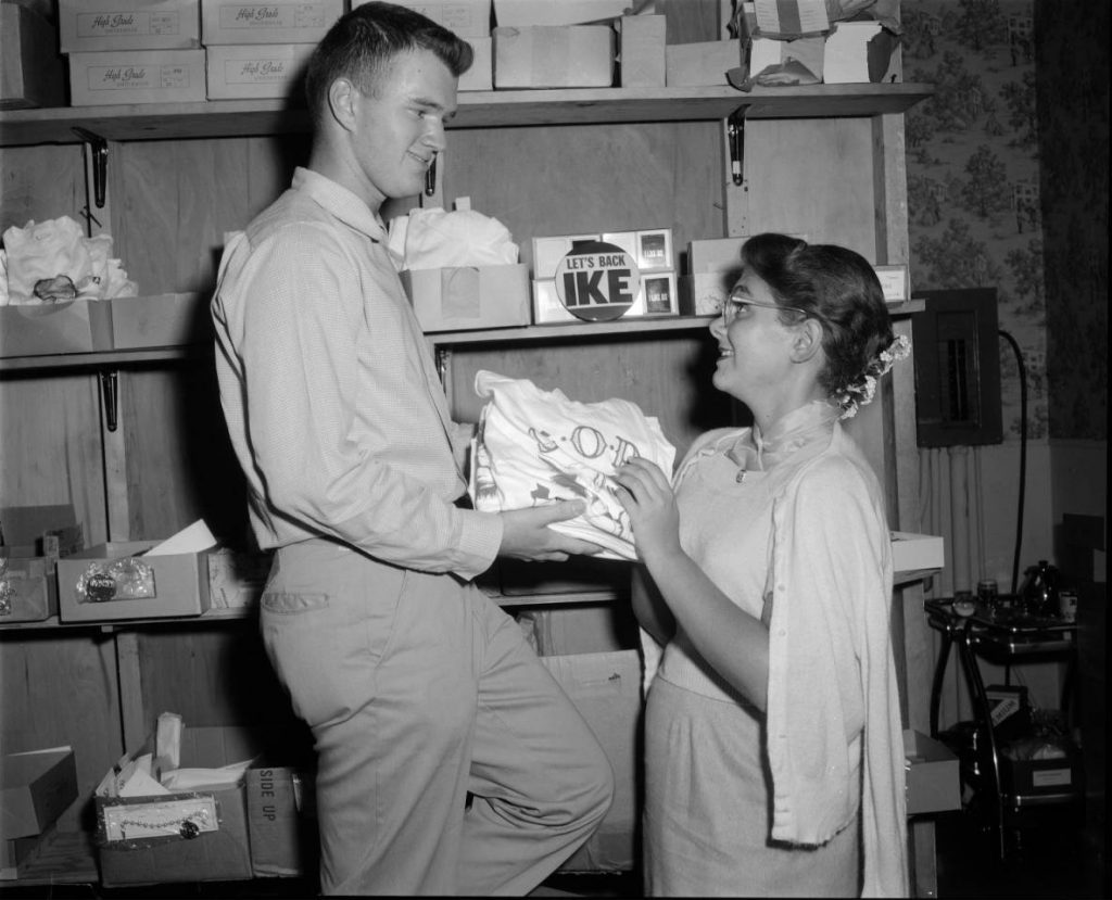 A young man and a woman are seen handling GOP and Dwight D. Eisenhower merchandise in 1956. The young man appears to be handing the young woman a stack of GOP shirts. Behind them is a large "Let's Back Ike" button and other shirts and promotional materials.