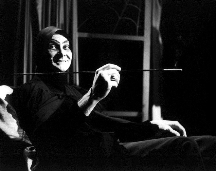 A man in ghoulish clothing sits holding a very long cigarette holder.