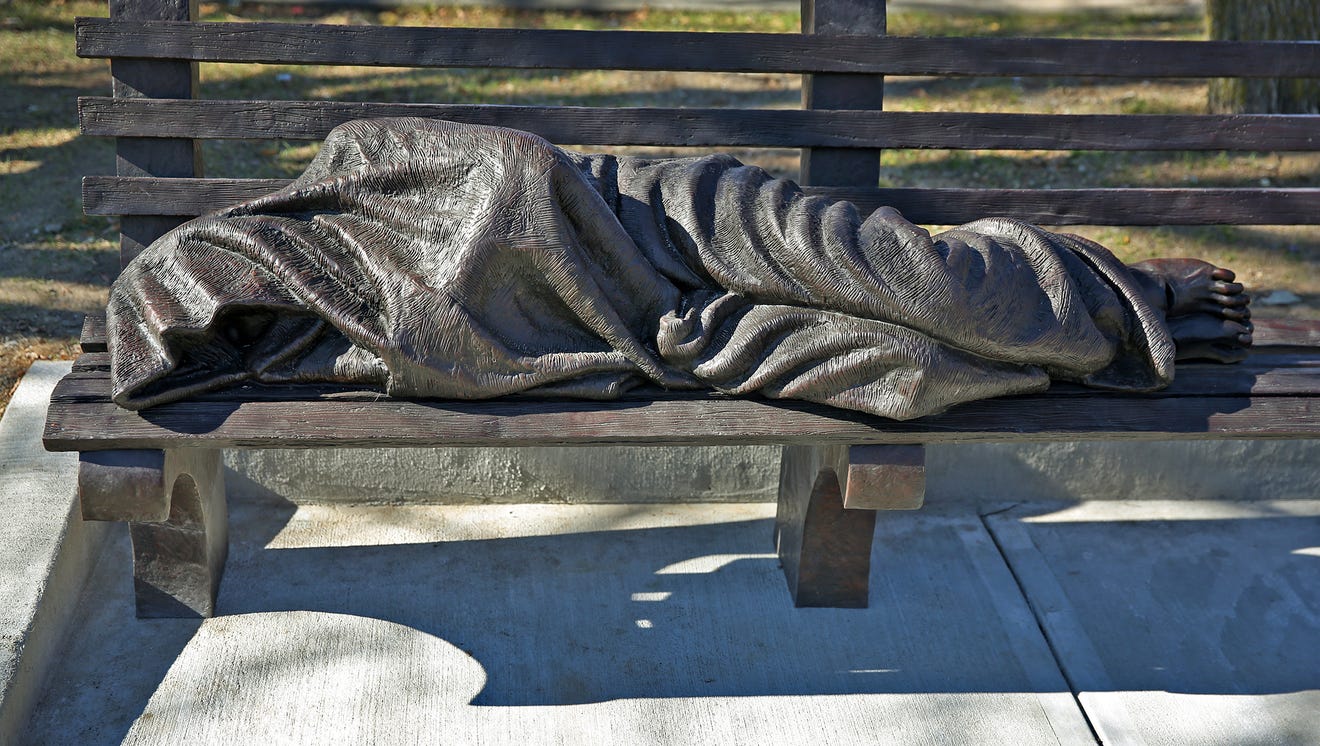 A sculpture of a covered person laying on a bench.