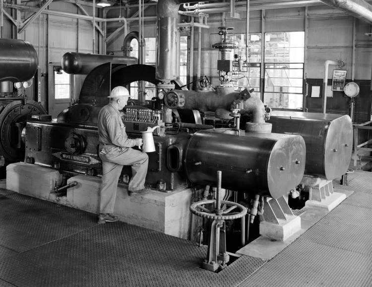 A man holds an oil can up to a large piece of machinery in a factory.
