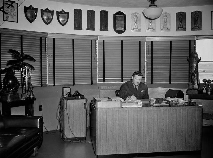 Colonel Roscoe Turner sits at a large desk. A line of plaques line the wall. 