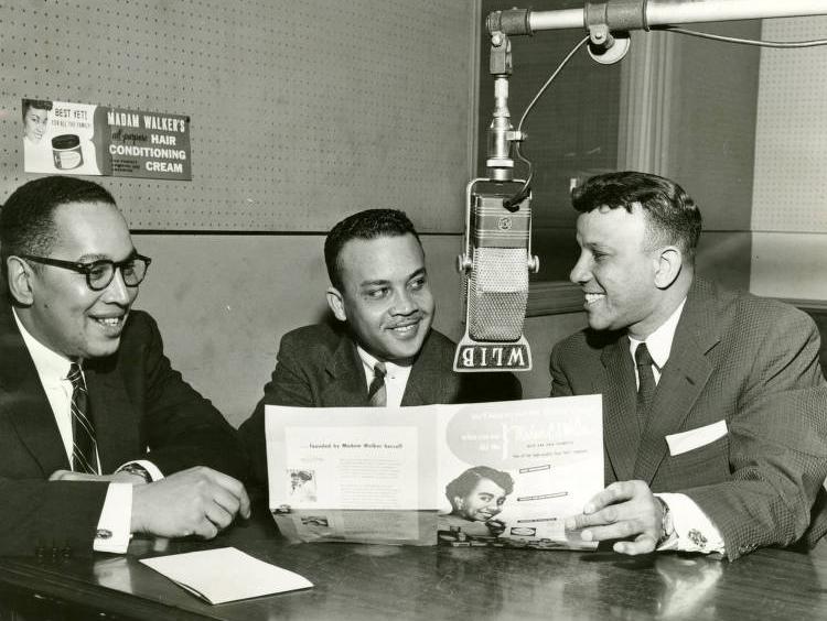 Three men are seated around a table. There is a microphone hanging from a boom in the center of the table.