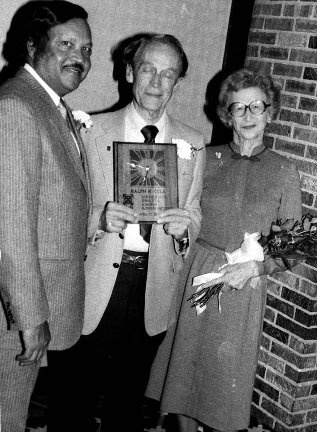 Two men and a woman are standing together. The man in the center is holding a plaque, and the woman is holding a sheaf of roses.