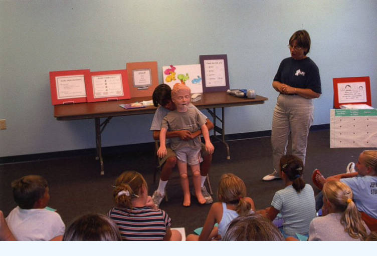 A child demonstrates the heimlich maneuver on a training manikin. An instructor and a group of children watch the demonstration.