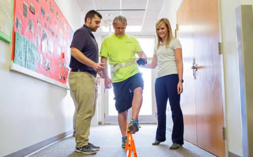 Two people stand on either side of another person who is attempting to walk over cones.