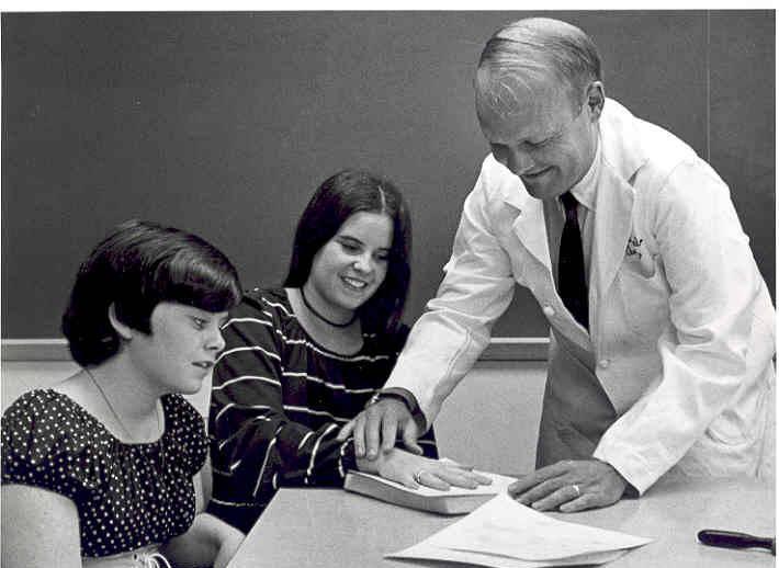 A person in a lab coat presses a young woman's hand to a pad of paper. Another young woman sits next to the first and watches.