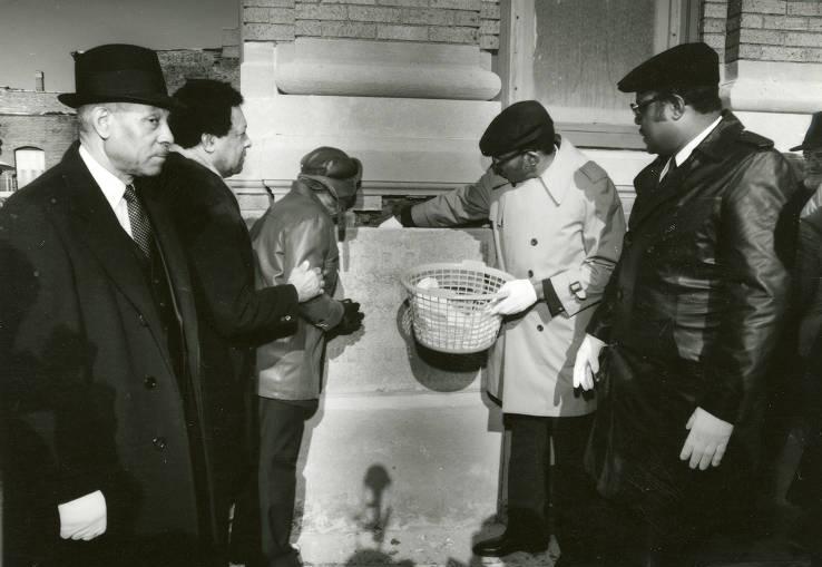 A group of men stand next to the corner of a buidling. Two men are either placing item in or remove items from a time capsule in the cornerstone.