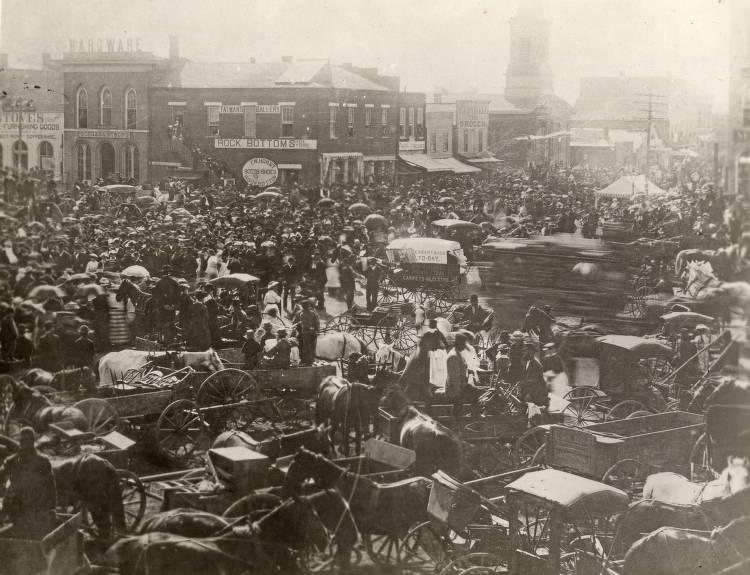 A crowd of people fill a large public square. Buildings line the square in the background and wagons fill the foreground.