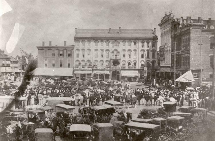 A large four-story hotel building is surrounded by other buildings. A large public square filled with people and wagons is in front of the buildings. 