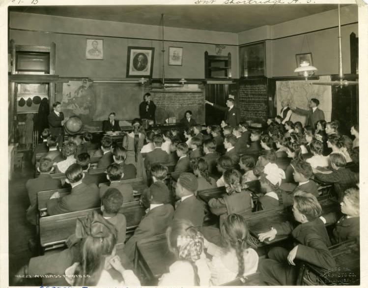 Students sit at desks in a classroom. A few students are at the front of the room presenting something.
