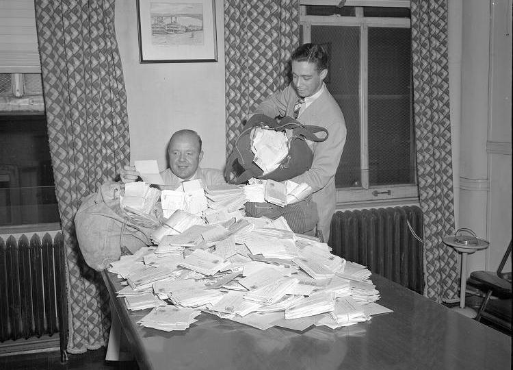 A man seated at a table sorts through a large pile of letters being dumped onto the table by another man.