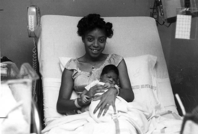 A woman sits in a hospital bed, holding a newborn infant.