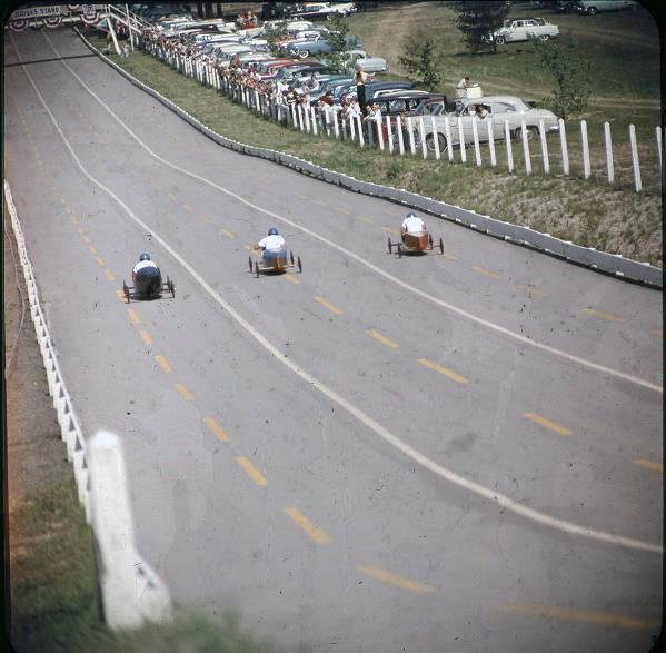 Three racers in soap box cars roll down a paved hill. 