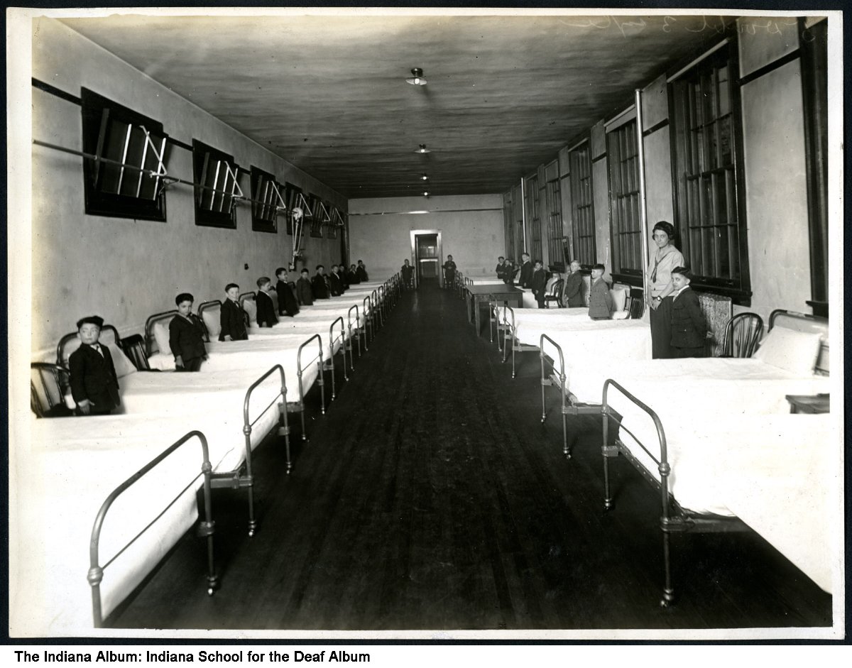 Rows of beds line two walls in a large room. Young boys stand next to each bed.
