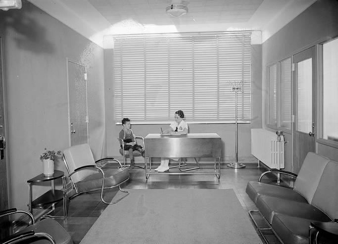 A nurse sits behind a desk. A young boy sits in a chair next to her.