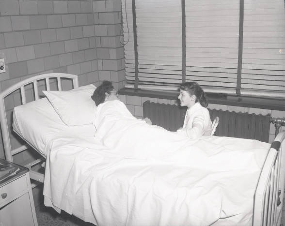 A young woman sits in a chair next to a woman in a hospital bed. 