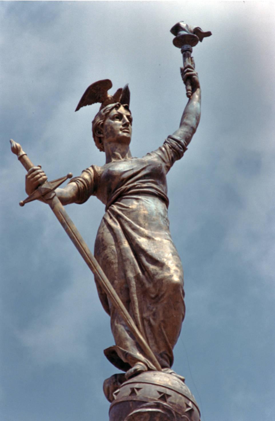 A bronze statue of a woman holding a large sword in one hand and a torch in the other. A bronze bird is perched on her head.