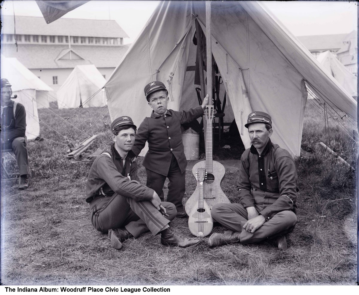 Three men pose with instruments outside of a tent. 
