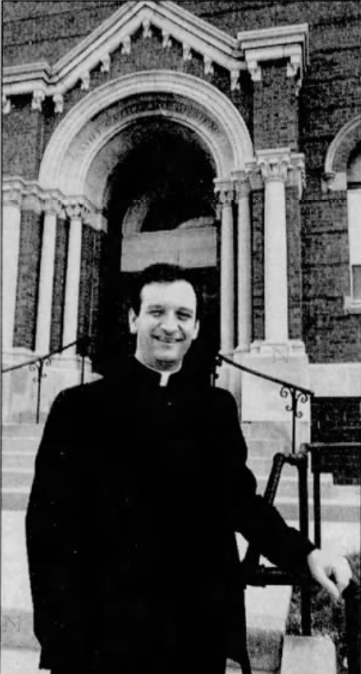 A man in clergy garb stand in front of the entrance to a church.